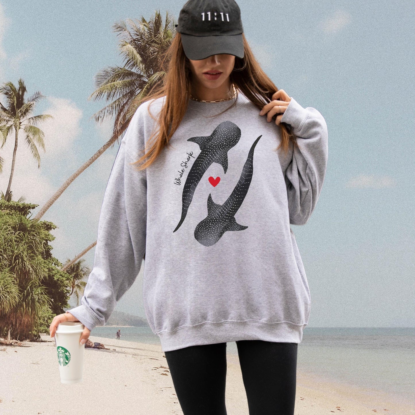 A Woman on the beach in a dad hat and grey sweatshirt with a Whale shark design